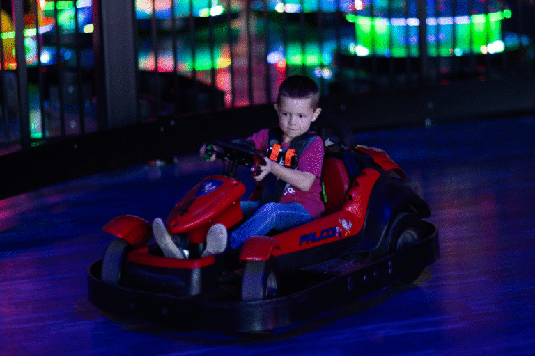 A young boy joyfully rides a go-kart at The Rush Funplex carnival, surrounded by colorful attractions and excitement.