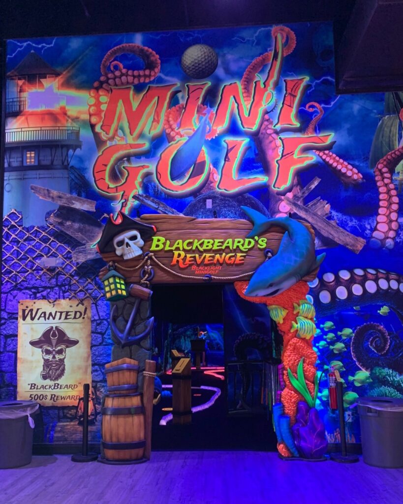 Inside The Rush Funplex, a dark room with neon lights surrounds a lively indoor golf course.