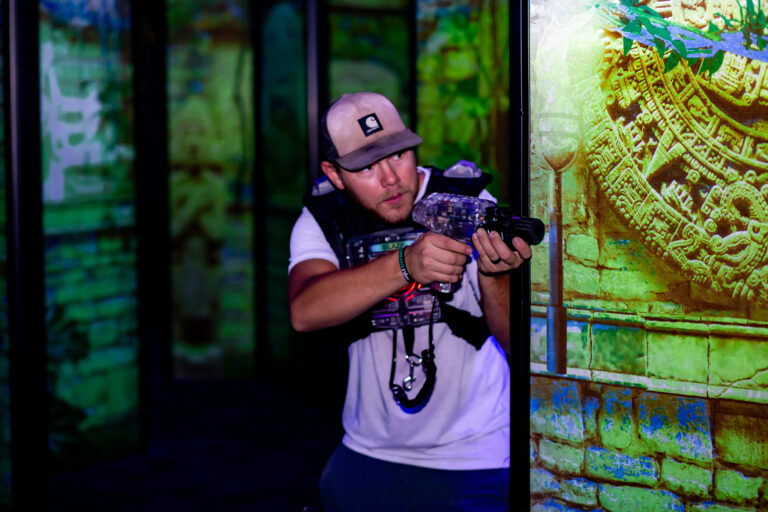 A man grips a gun while standing before a wall at The Rush Funplex, evoking a sense of suspense or intensity.